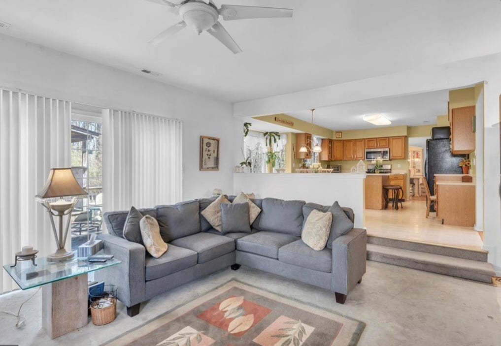 Modern living room with gray sectional sofa, wooden kitchen visible in background, ceiling fan, and large windows with white curtains