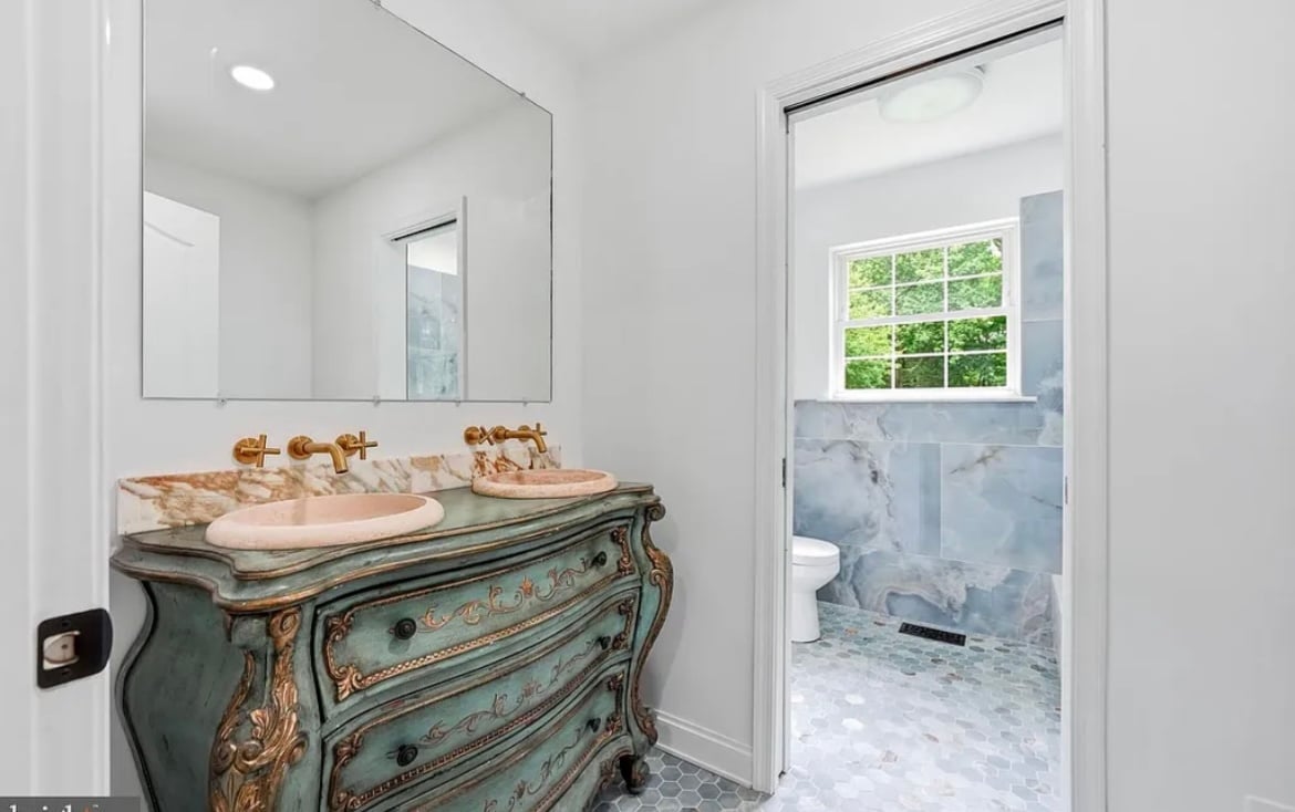 Bright bathroom with vintage green vanity, marble-look sinks, large mirror, and adjacent tiled shower with window