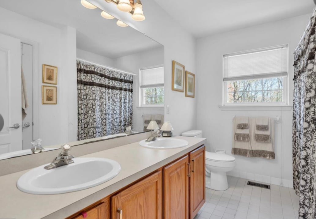 Bright, clean bathroom with wooden vanity, double sink, white countertop, and shower-tub combo behind patterned curtains