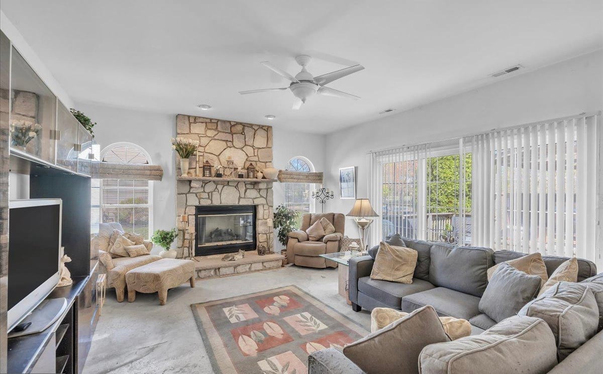 Spacious living room with stone fireplace, beige and gray furniture, area rug, ceiling fan, and glass doors opening to outdoor patio