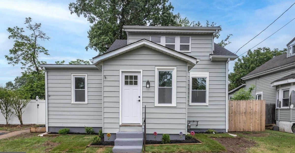 A small gray and white house with vinyl siding, white trim, and a front porch, surrounded by landscaping and trees on a sunny day.