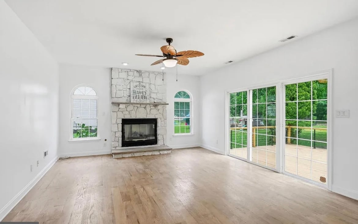 Empty modern living room with wood ceiling fan, fireplace, hardwood floors, large windows, and glass patio doors