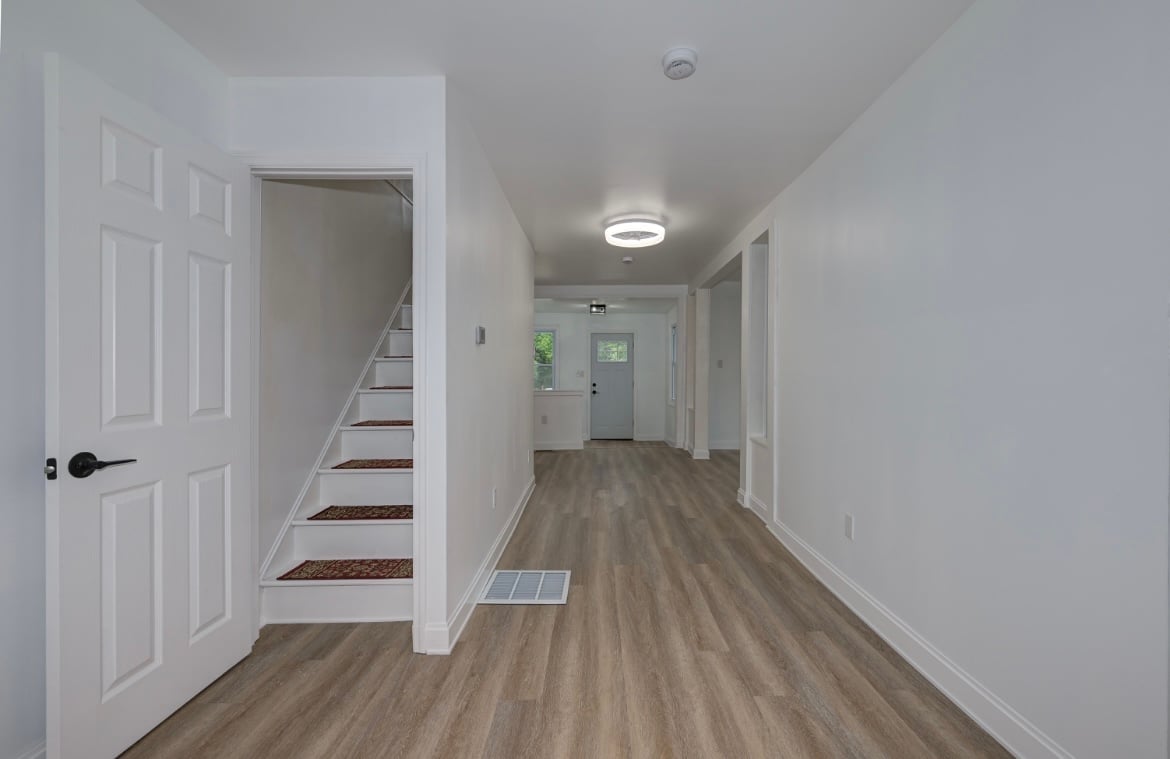 Interior hallway with white walls, wood plank flooring, LED ceiling light, staircase entry, and white panel doors