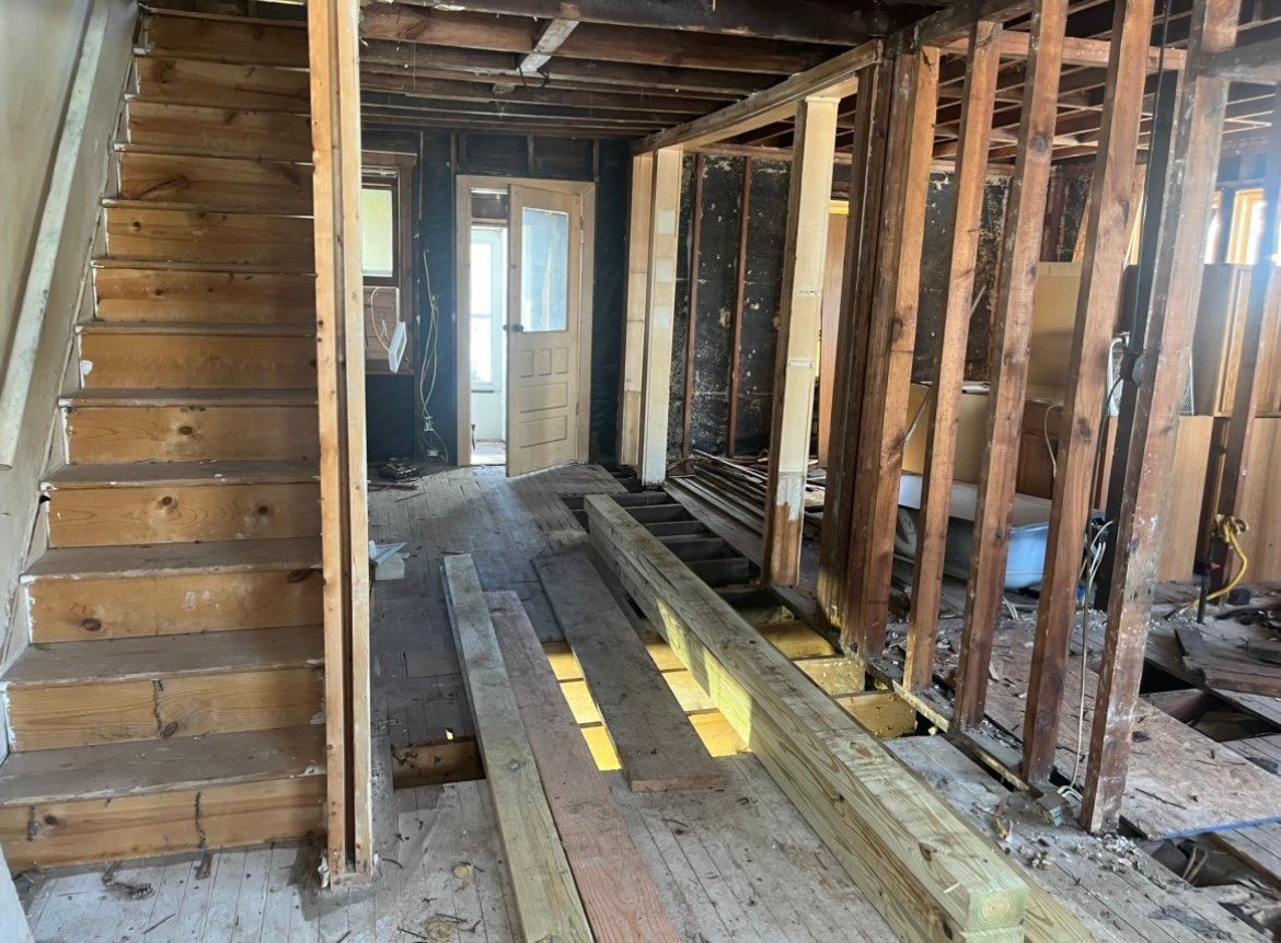 Interior of a house under renovation showing exposed wooden beams, framing, and subflooring with yellow safety markings