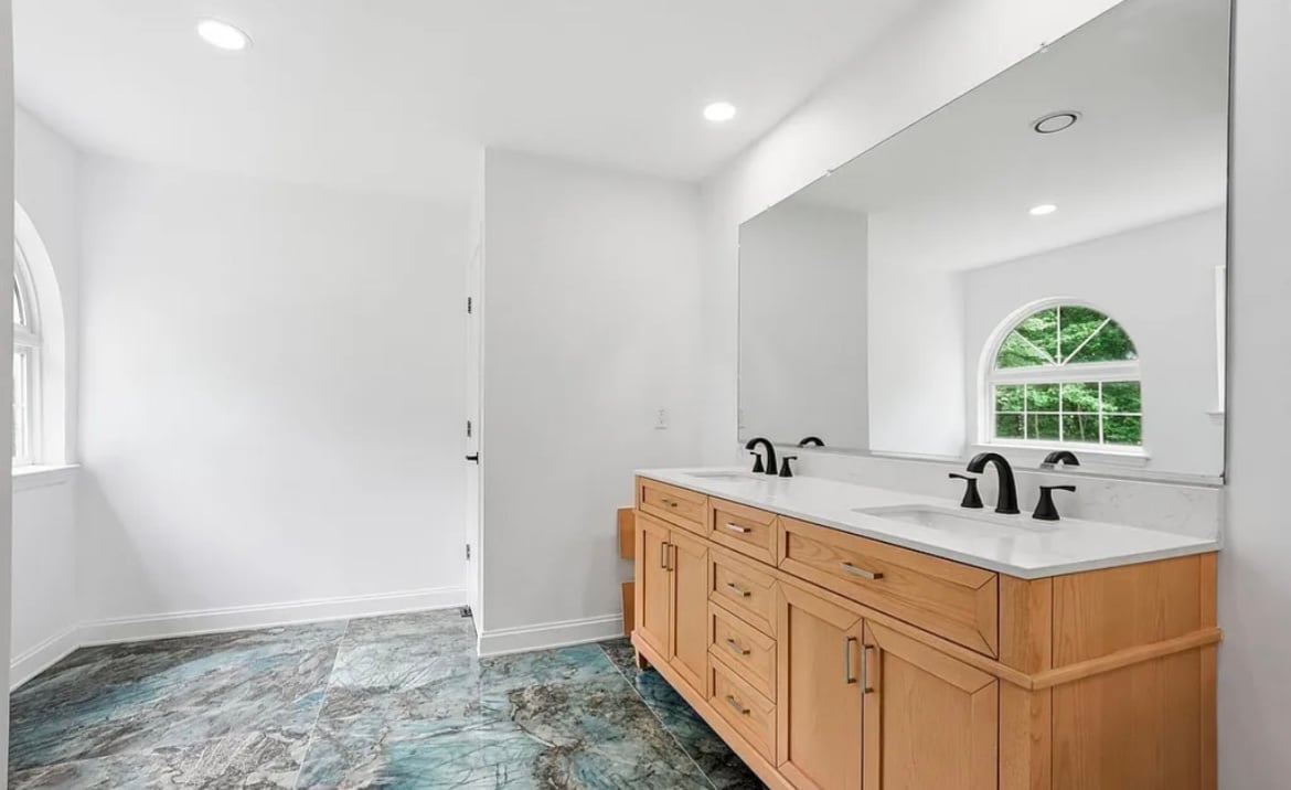Modern bathroom with wooden double vanity, white countertops, large mirror, and arched window overlooking landscaped grounds