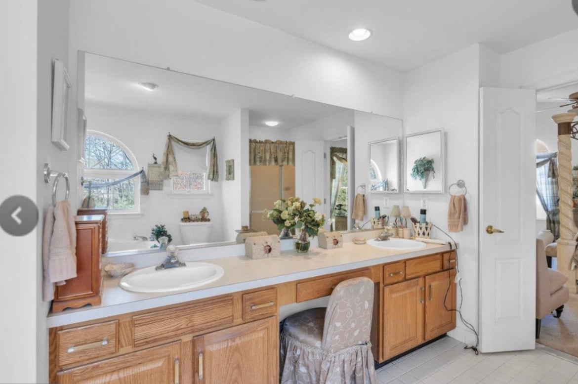 Bright master bathroom with dual wooden vanities, white countertops, large mirror, and decorative accessories on shelves above sink